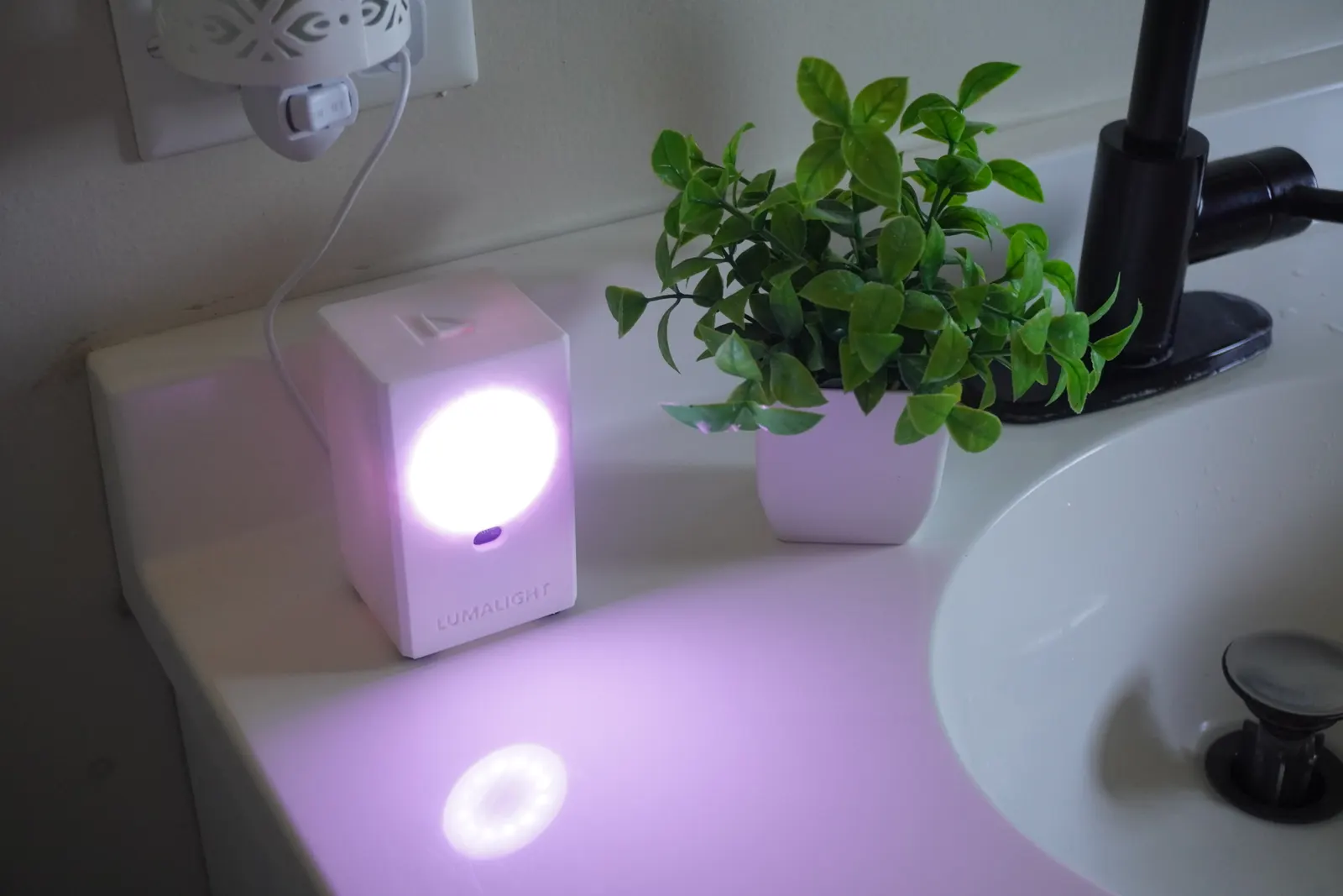 LumaLight on a bathroom counter beside a potted plant, its ring lit pale purple and casting a circle of light on the counter